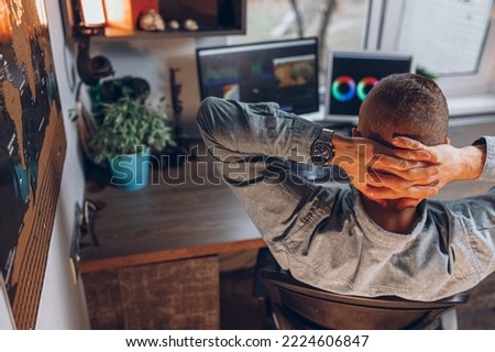 High angle back view of male video maker relaxing with hands behind head during video editing on computer monitors while working in home office 