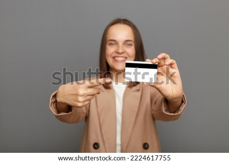 Photo of satisfied brown-haired woman wearing jacket pointing at plastic credit card, recommend bank or shopping discounts, looking at camera with toothy smile, posing isolated over gray background.