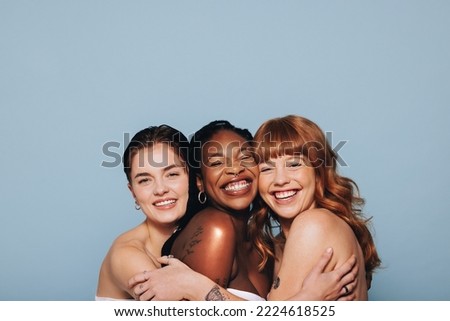 Three happy women with different skin tones smiling and embracing each other in a studio. Group of diverse women feeling comfortable in their natural skin. Body positive young women standing together. Royalty-Free Stock Photo #2224618525