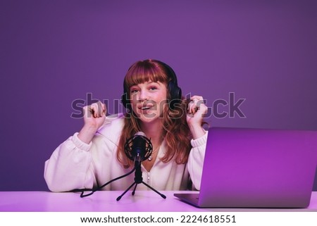 Happy young podcaster speaking into a microphone in a recording studio. Young woman hosting a live audio broadcast against a purple background. Woman creating content for her internet podcast.