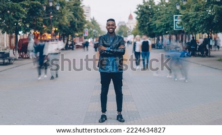 Portrait of happy African American guy wearing stylish jeans and leather jacket standing alone in street downtown, smiling and looking at camera with crowd moving by. Royalty-Free Stock Photo #2224634827