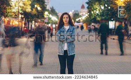 Portrait of stylish young lady tired of usual haste standing in the street among whizzing people and looking at camera. Time, youth and modern society concept. Royalty-Free Stock Photo #2224634845