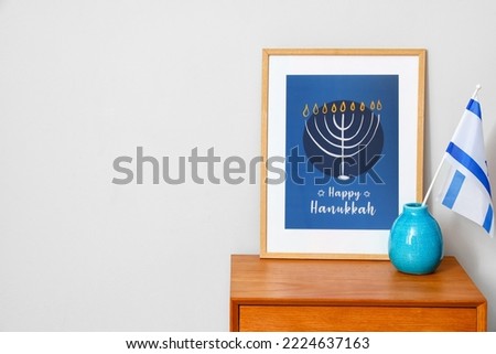 Frame with text HAPPY HANUKKAH and flag of Israel on table near light wall