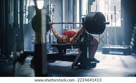 A Professional Bodybuilder Training by Lifting a Heavy Barbell In a Dark Gym. Female Energetic Athlete Doing Core Exercise with Bench Press Workout in an Empty Gym Royalty-Free Stock Photo #2224641413