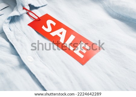 Close-up red tag with advertising text Sale hanging on light-colored shirt in clothing store. Black Friday concept.