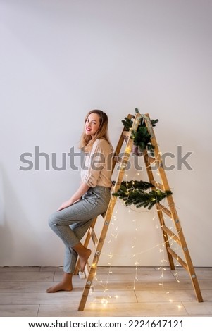 Portrait of modern middle aged woman at wooden ladder, decorated with fir branches, garland for Christmas. Place for text, light wall. Scandinavian style in the interior of the house for holiday.