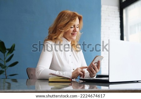 Beautiful business woman blonde in a blouse sits at a desk in front of an open laptop, holds a mobile phone in her hands and answers messages. Horizontal photo
