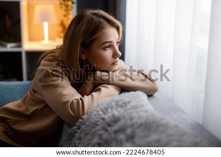 Side view of sad caucasian woman in casual hoodie leaning with head on hands while sitting on couch and looking at window. Boring at home. Royalty-Free Stock Photo #2224678405