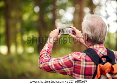 Retired Senior Man Hiking In Woodland Countryside Taking Photo With Mobile Phone
