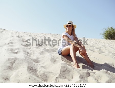 A young woman in a white t-shirt and hat sits on the sand in the desert. The woman looks into the distance. Brown tint. Woman portrait