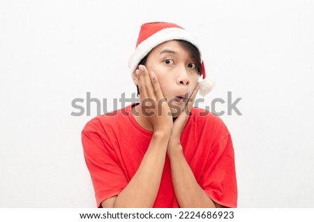 expressive happy young asian man in christmas santa theme clothes wit shocked, surprised and amazed face expression. wow expression man in red santa hat. christmas model concept for sale and discount.
