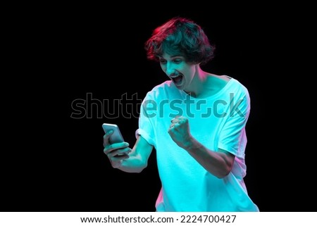 Portrait of young man with mustache looking at phone with happy excitement isolated over black background in neon light. Concept of youth, lifestyle, emotions, facial expression. Copy space for ad