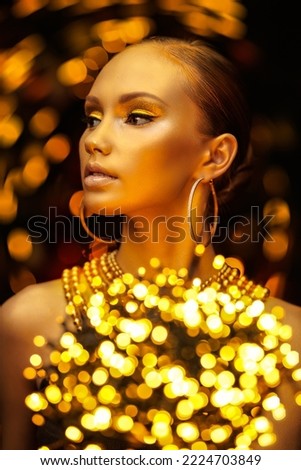 Golden Christmas girl. Stunning woman with golden make-up and skin wearing golden earrings and necklace on a dark background with sparkling golden lights around. Holiday luxury look. 