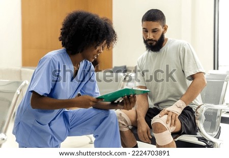 African black nurse woman asking injured informations from young Caucasian patient man in hospital, female afro hair nurse in blue hospital uniform checking beard man patient about injured wound Royalty-Free Stock Photo #2224703891