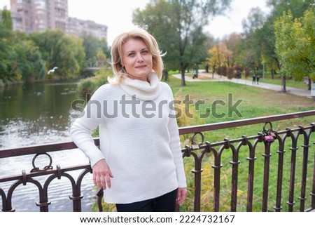 Blonde woman in white sweater against walk in city park on a bridge in autumn. Dreamy European lady against the backdrop of reaver in city park. Middle-aged woman on a walk in the city.