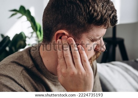 Close-up of unhealthy young caucasian man 30s touching ear, suffering from sudden throbbing ear ache, looking aside. Upset bearded male feeling unwell sitting on couch in living room at home Royalty-Free Stock Photo #2224735193