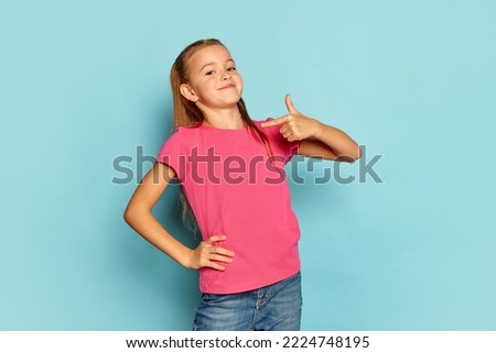 Portrait of little beautiful girl, child in pink T-shirt posing isolated over blue studio background. Proudly pointing at herself. Concept of childhood, emotions, facial expression, lifestyle Royalty-Free Stock Photo #2224748195