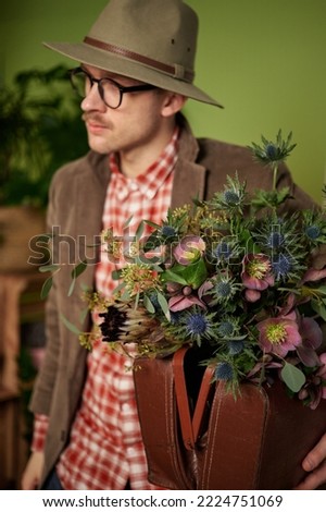 Attractive vintage styled male person in eyeglasses, velvet jacket and brown hat staying with flower bouquet and briefcase in flower store. High quality vertical image