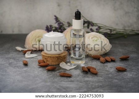 Natural homemade face serum and cream. Transparent glass bottle and white plastic cream container. Grey marble background with flowers and almonds. 