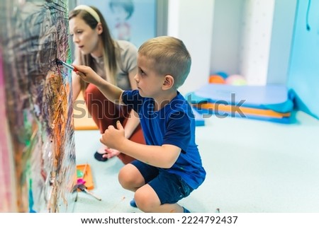 Caucasian little boy in blue clothes holding a brush and painting on the cellophane in the nursery, teacher in the background. High quality photo