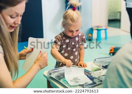 blond cute girl painting with water and paints with her teacher in the nursery, medium shot. High quality photo