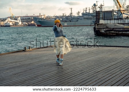 Outdoors fashion portrait of a beautiful middle aged woman walking on the beach. Marine background. Dressed in a stylish warm blue sweater, yellow skirt and beret.