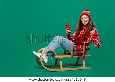 Full body merry young woman wearing red warm cozy knitted sweater hat posing sledding use mobile cell phone show thumb up isolated on plain dark green background. Happy New Year 2023 holiday concept