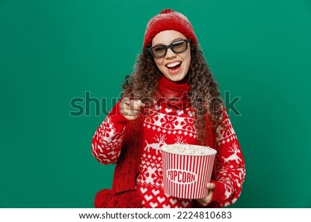 Young woman in red knitted sweater hat 3d glasses watch movie film hold bucket of popcorn in cinema point finger camera on you isolated on plain green background Happy New Year 2023 holiday concept