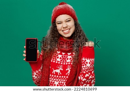 Merry young woman wearing red warm cozy sweater hat posing hold in hand use mobile cell phone blank screen workspace area isolated on plain dark green background. Happy New Year 2023 holiday concept