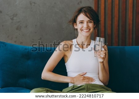 Young woman wears white tank shirt hold drink clear fresh pure still water from transparent glass put hand belly sit on blue sofa stay home flat rest relax spend free spare time in living room indoors