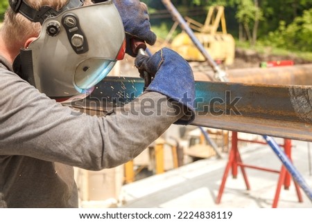 A professional welder in a protective suit and mask produces the connection of pipes with the help of a welding machine on the construction site. Royalty-Free Stock Photo #2224838119