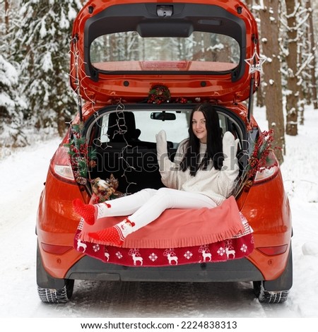 The trunk of the car is decorated with Christmas decorations. A beautiful cheerful girl is sitting in the trunk of a car, laughing and waving her hands. Car on the road in the winter forest. 