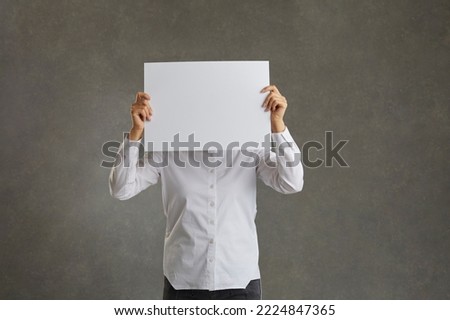 Man holds a blank white sheet of paper in front of his face, free space. The man is holding a sheet of paper horizontally in front of his face, against a black background.
