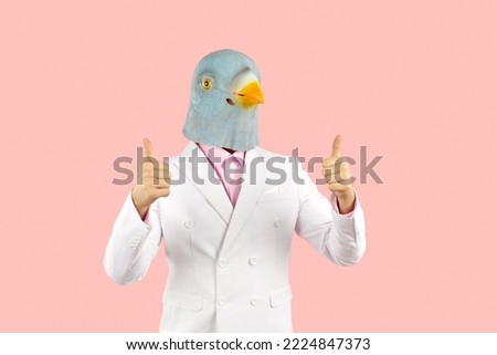 Content man with funny bird face shows thumbs up with both hands. Surreal shot of supportive guy wearing white suit and bizarre pigeon mask standing on pink color background and showing Great Job sign