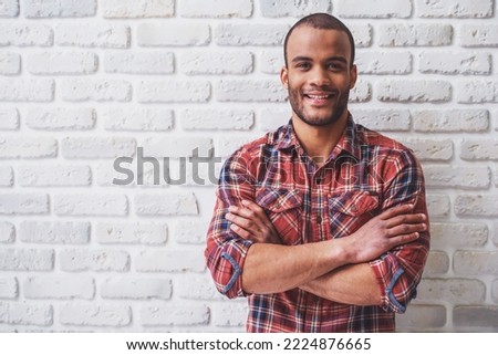 Handsome Afro American man is looking at camera and smiling, standing with crossed arms against white brick wall