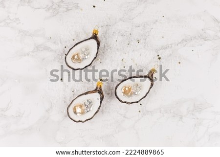 Three Christmas tree baubles in the shape of an oyster are lying on a marble table. Glitter and stars decorate the picture.