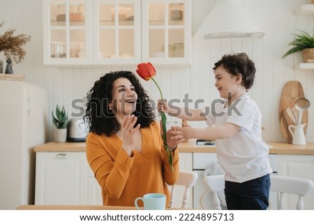 little curly boy in a white T-shirt and blue pants in kitchen gives his curly beautiful mother a tulip on mother's Day. cheerful boy dares his mother a flower, , they sit in the kitchen together.