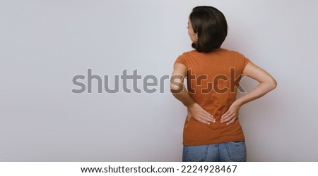 Spinal cord problems on woman's back. Chronic back pain. Young brunette woman is holding her lower back, while standing and suffering from unbearable and severe pain. Spine osteoporosis. Royalty-Free Stock Photo #2224928467