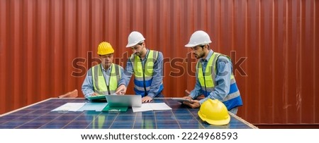 Project manager pointing at  shipping schedule on laptop computer, explain to contractor  about planning for next shipment. They use solar cell panel as a table. A large container is in the background