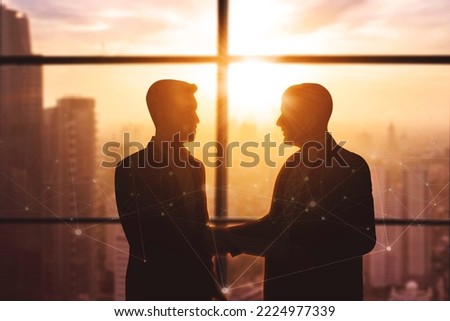 Silhouette of two businessmen handshaking together while standing near window with dusk sunlight background
