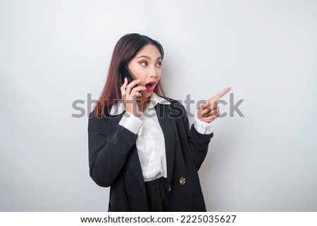 Shocked Asian businesswoman wearing a black suit pointing at the copy space beside her while talking on her phone, isolated by a white background