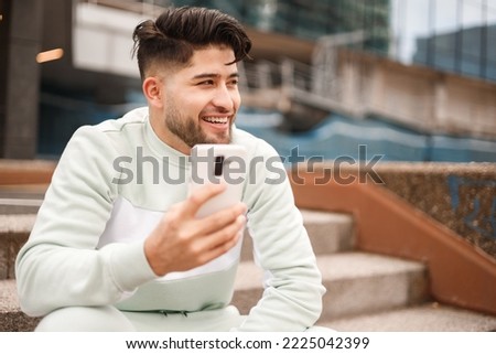 happy hispanic young man in sportswear using cellphone in urban scenario, big closeup view