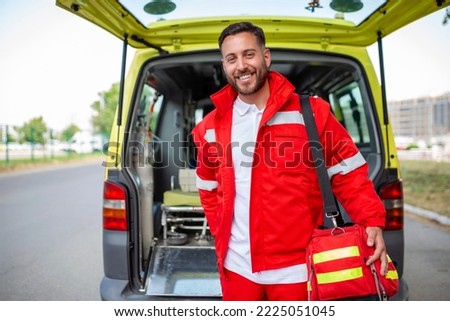Young man , a paramedic, standing at the rear of an ambulance, by the open doors. He is looking at the camera with a confident expression, smiling, carrying a medical trauma bag on his shoulder. Royalty-Free Stock Photo #2225051045
