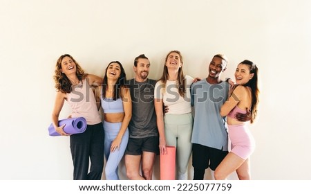 Group of happy fitness friends laughing while standing against a wall with yoga mats. Multicultural people attending a yoga class together. Sporty people working out in a community fitness studio. Royalty-Free Stock Photo #2225077759
