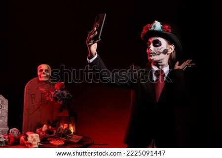Goddess of death taking pictures on mobile phone, wearing dios de los muertos halloween costume and make up. Woman capturing image being dressed as la cavalera catrina over black background.