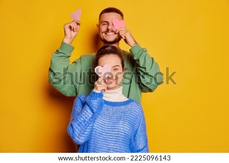 Portrait of beautiful young couple, man and woman posing with paper hearts isolated over yellow background. Happy celebration. Concept of love, relationship, Valentine's Day, emotions, lifestyle