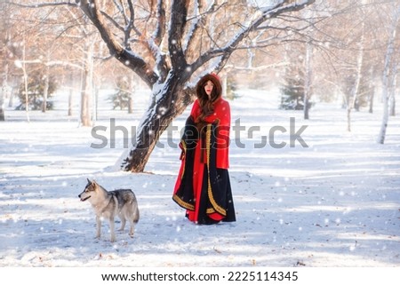 A young woman in a red hooded raincoat with a gray husky dog on the background of a winter forest and snowfall.