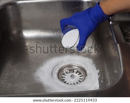 Gloved hand putting baking soda on drain in kitchen sink from glass jar. Close up. Eco friendly house cleaning concept. Royalty-Free Stock Photo #2225119433