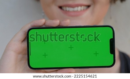 A pretty girl with a sweet smile is holding a modern mobile phone horizontally with a green screen facing camera. Use green screen for copy space closeup. Chroma key mock-up on smartphone. 