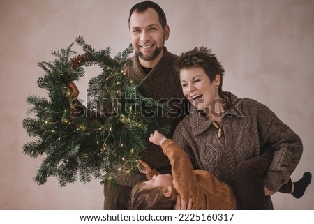 Happy family in knitted sweaters with a New Year's wreath and a garland on a beige grunge background. Fashionable Christmas handmade style. Mom, dad and son in hipster style